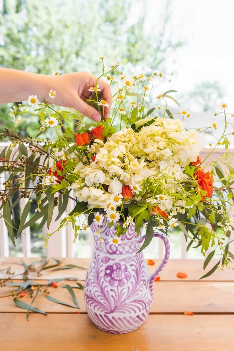 A person places chamomile blossoms in a bouquet of eucalyptus, roses, and hydrangeas in a purple and white vase.