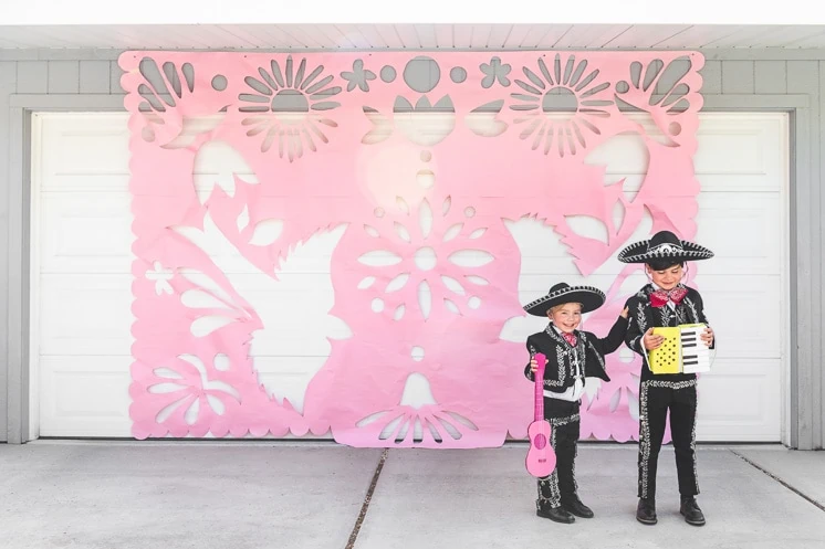 Two little boys dressed in Mariachi outfits holding cardboard instruments stand in front of a giant pink papel picado decoration