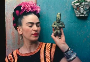 Photograph of Frida Kahlo holding a small carved idol. She's wearing a black and orange woven top and has flowers and ribbons braided into her hair, and she's standing against a turquoise wall.