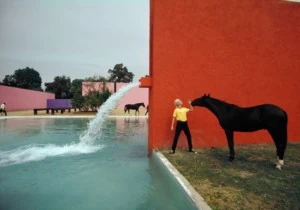 A woman holds a black horse's bridal in front of a tomato-colored wall at Luis Barragán's Cuadra San Cristóbal stables. There's also a flat blue pool and pink and purple structures in the background.
