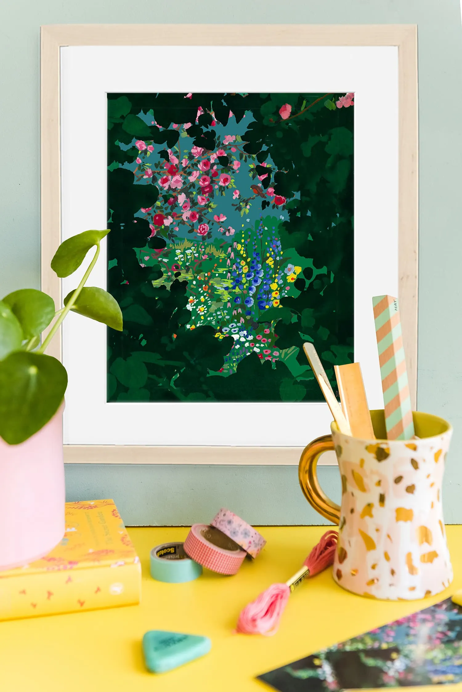 A print of flowers in a green secret garden tunnel hangs on the wall in front of a plant and some craft supplies