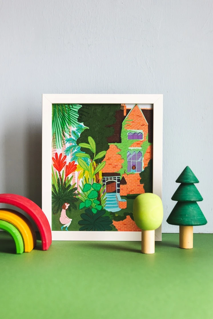 An art print of girls inside and outside a brick building covered with vines and surrounded by trees and flowers. Next to the print are wooden toys shaped like trees and a rainbow.