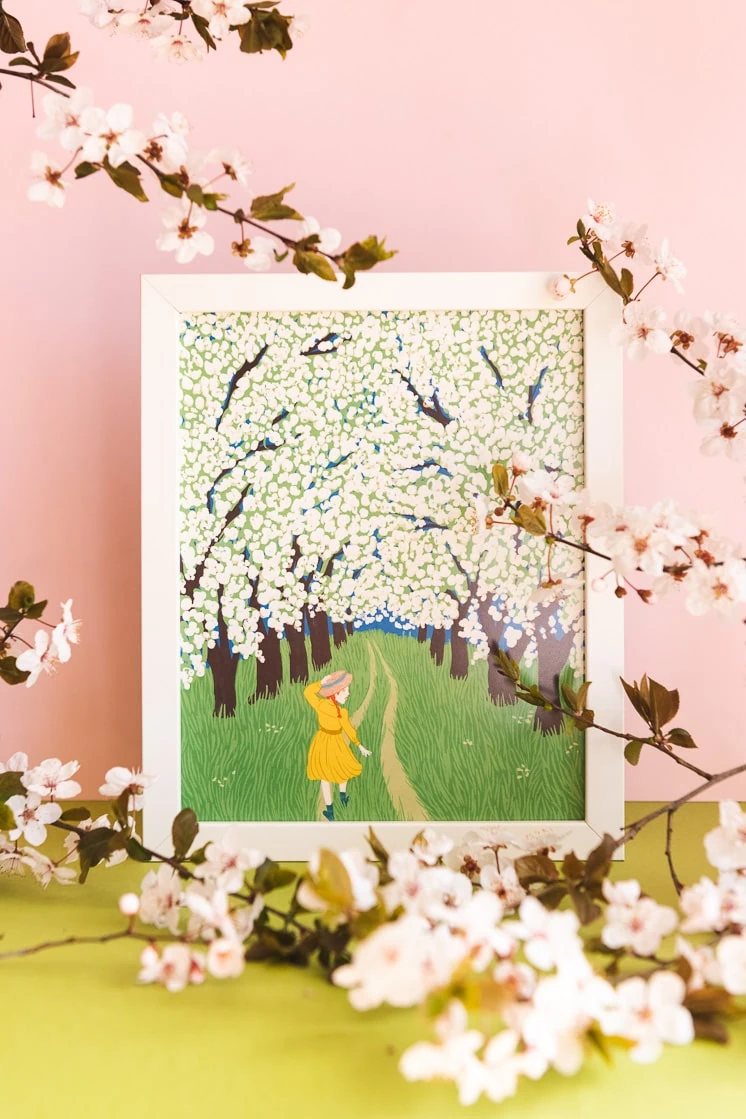A print of Anne Shirley from Anne of Green Gables walking through a corridor of blossoms against a pink background with real blossoms in front.