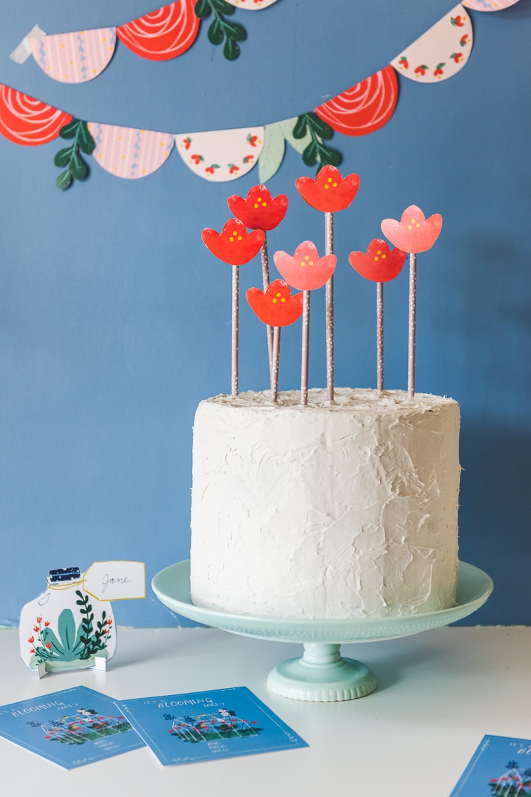 Garden-themed paper bunting swoops across a cornflower blue background. Terrarium-shaped place markers and blue invitations with a garden illustration sit on the table next to a white frosted cake with red and pink illustrated floral cake toppers.