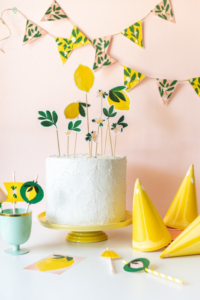 Lemon-themed bunting swoops across a pink backdrop. In the foreground, straw toppers, invitations, and lemon-slice party hats sit in front of a white-frosted cake with lemon and lemon blossom cake toppers.