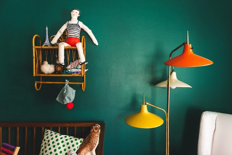 An interior shot featuring a painted green wall with a brightly colored lamp and a toy doll perched on a wicker shelf.