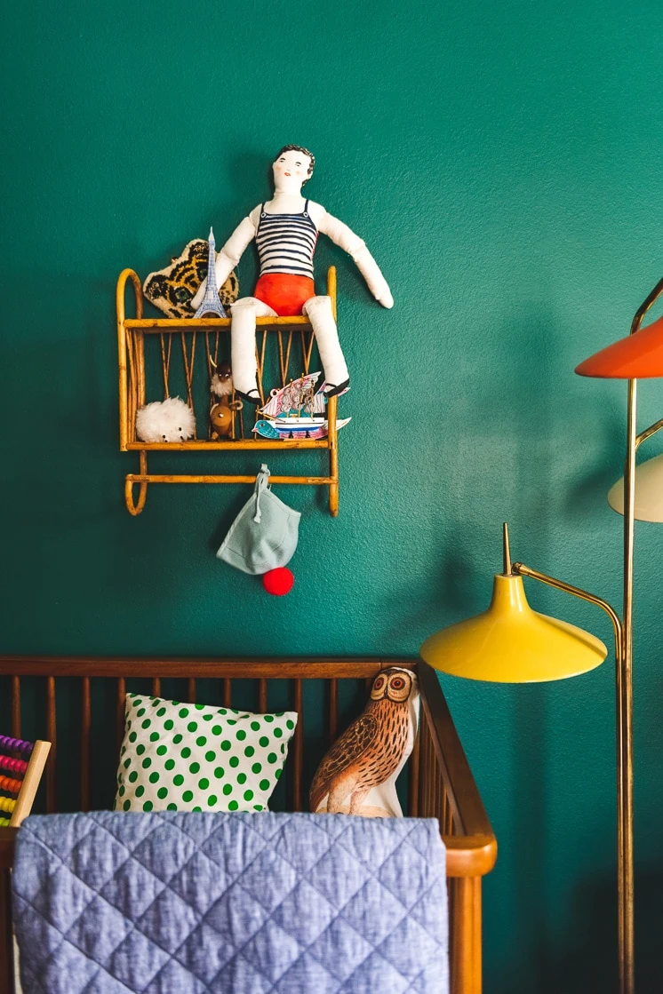 Interior shot of a child's room with a green wall, a wicker shelf with a toy on it, and a crib. The crib has a denim-colored quilt hanging over the side. 