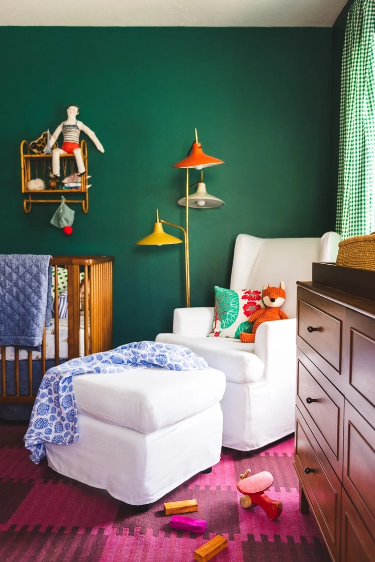 Interior shot of a child's room. Walls are green, A pink checkerboard rug is on the floor. A white rocking chair is central in the image.