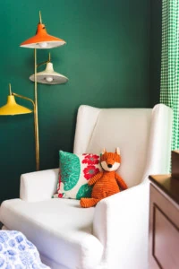 A white rocking chair against a green wall with a colorful lamp in the background. An orange stuffed fox and a pillow are on the chair.