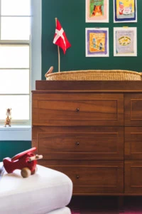 Interior shot of a nursery. In the foreground, a red toy airplane rests on a white ottoman. In the background is a wooden dresser with a small swedish flag on top and some illustrations on the wall.