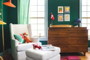 Interior shot of a green nursery. In the foreground is a white rocking chair with a few pillows, toys, and books on it and in the background is a wooden dresser.