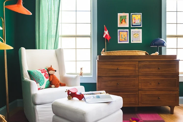 Interior shot of a green nursery. In the foreground is a white rocking chair with a few pillows, toys, and books on it and in the background is a wooden dresser.