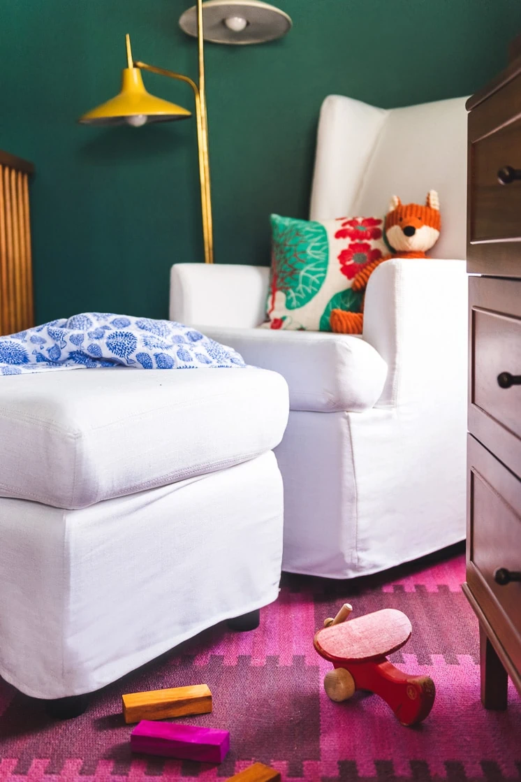 A white rocking chair against a green wall with a colorful lamp in the background. An orange stuffed fox and a pillow are on the chair. The floor is covered by a magenta checkerboard rug with a few wooden cars and an airplane on it.