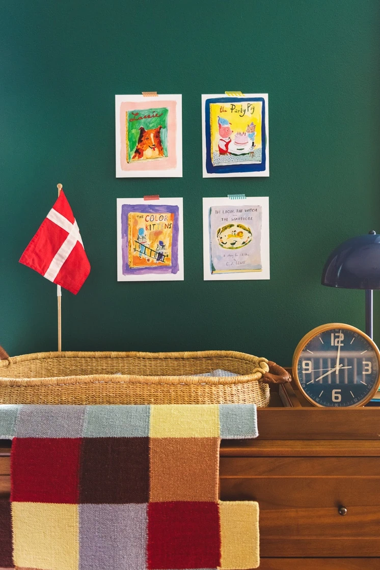 A wooden dresser with a clock, changing basket, Danish flag, and blanket on top. The wall has a few illustrations hanging on it. 