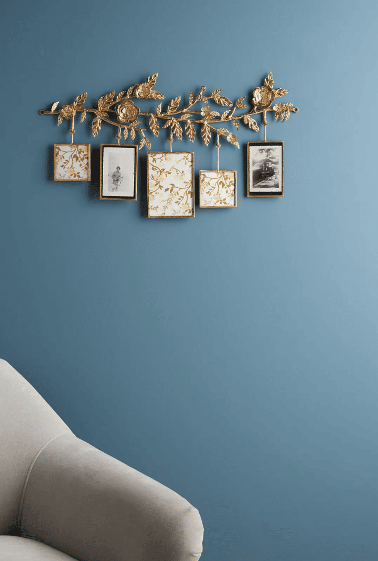 A golden metal branch with intricate metal flowers and leaves and five hanging rectangular picture frames below in a powder blue room.