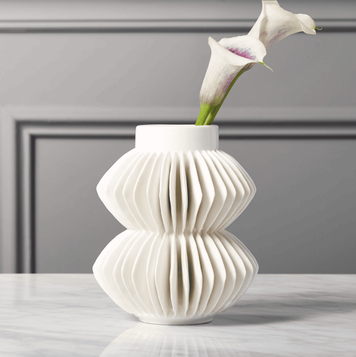 A white vase with ceramic, fan-like fins sits on a table. Two calla lilies are inside.