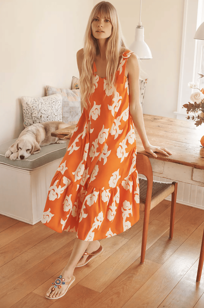 A woman leans against a chair in a light-filled room. She's wearing an orange sleeveless dress with a flounce at the bottom and abstract large white flowers printed on it. A dog lounges on a bench behind her. 