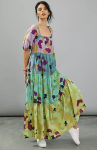 A woman wearing a tiered maxi dress with lilac, blue, aqua, and chartreuse floral tiers stands in a grey room