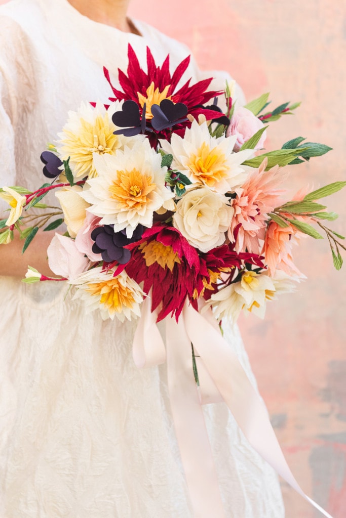 A bride in a white dress against a pink background holds a paper bouquet made of dahlias, roses, shamrocks, Mexican jasmine, and foliage.