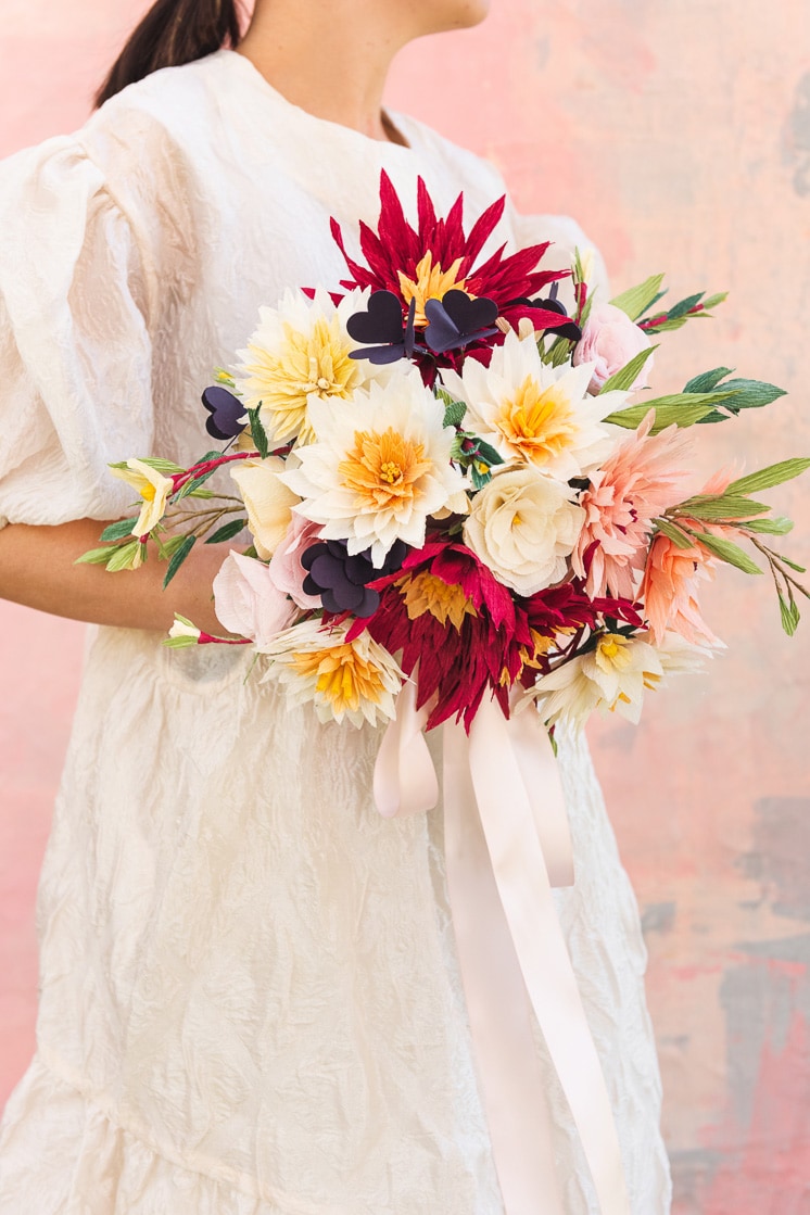 A bride in a white dress against a pink background holds a paper bouquet made of dahlias, roses, shamrocks, Mexican jasmine, and foliage.