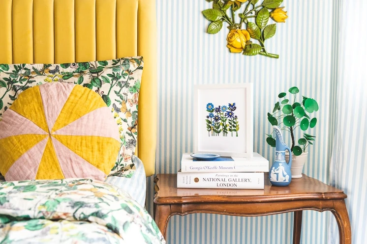 Field of Colored Flowers Papercut By Julie Marabelle is framed and perched on top of a stack of books next to a yellow bed and a blue and white striped wallpapered wall.