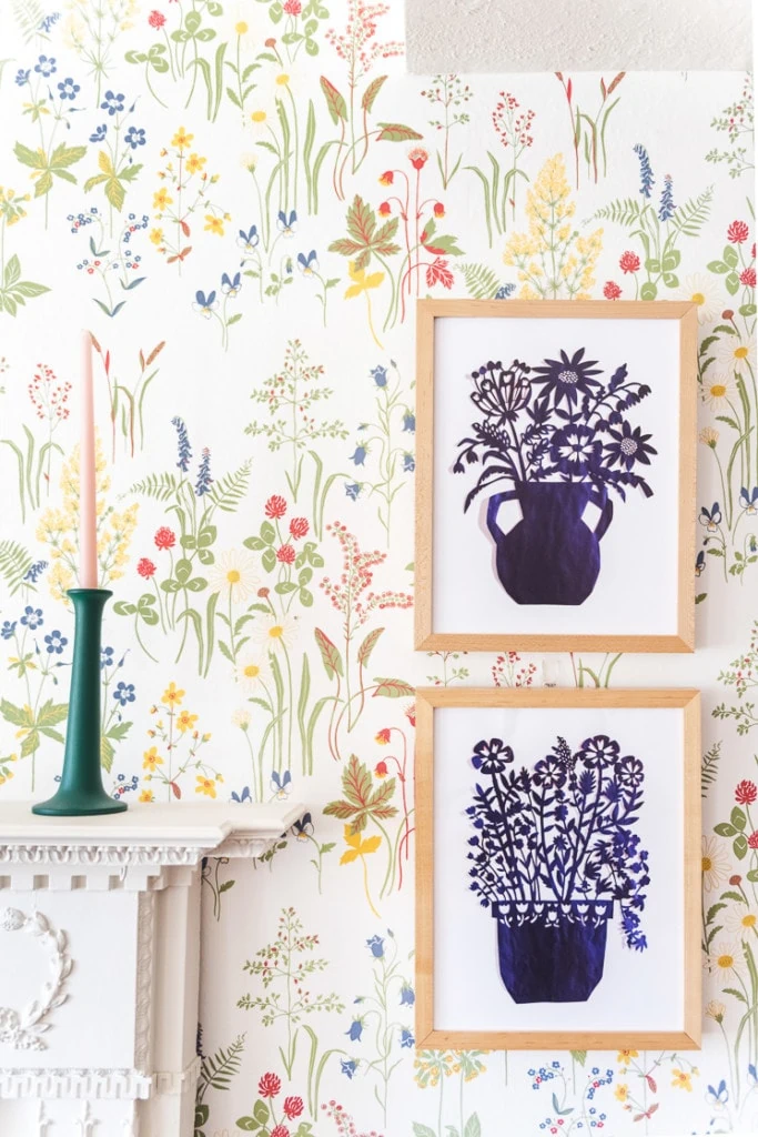 Two blue papercut art prints on a wildflower wallpaper wall next to a mantle with a green candlestick