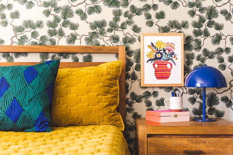 A framed paper-cut floral print on a spruce-themed wallpapered wall by a yellow bed and a blue lamp