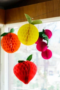 paper fruits in the shape of grapes, an orange, a grapefruit, and a strawberry hang in front of a window