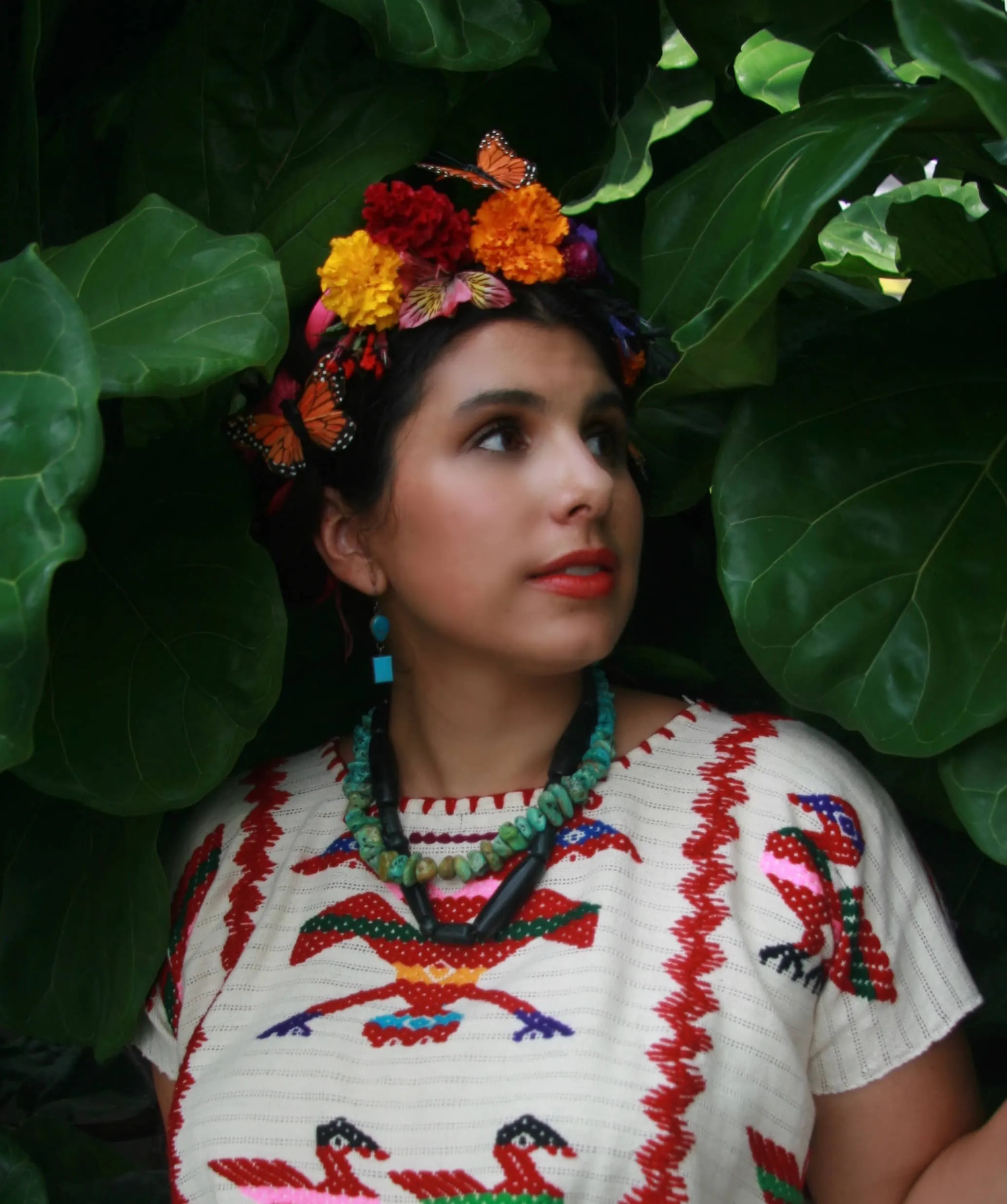 Michelle is wearing a traditional Mexican embroidered blouse and stone necklaces. She has flowers in her hair and is standing among leaves.