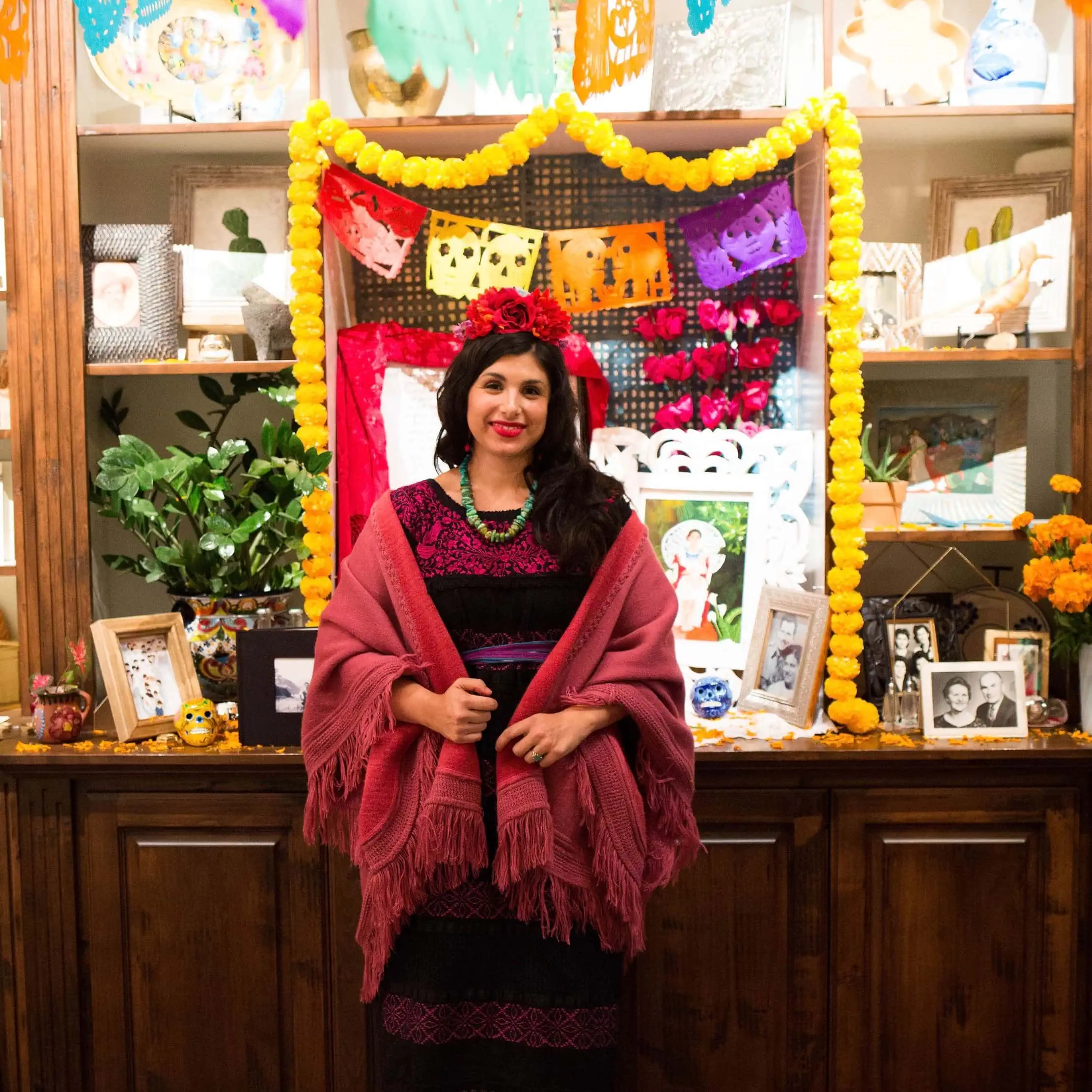 Michelle is wearing a red dress and rebozo. She has red flowers in her hair and she's standing in front of a Dia de los Muertos ofrenda. 