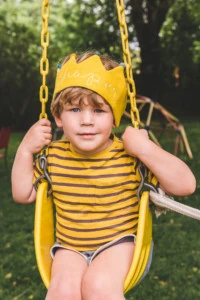 Jasper wears his Flower Lane crown and a yellow striped shirt on a yellow swing.