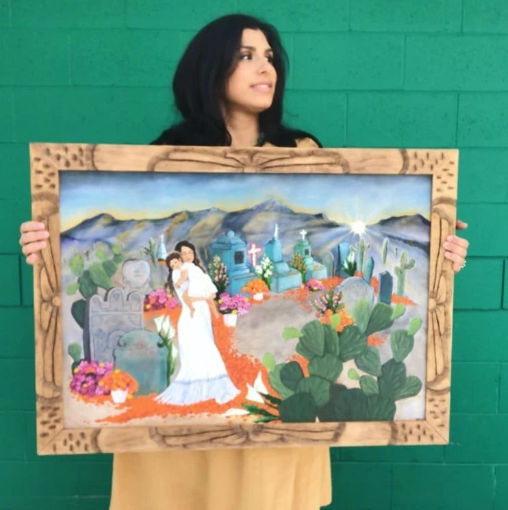 Michelle is holding up a painting of a woman holding a young girl in a Mexican cemetery surrounded by marigolds, nopales, and mountains. She's against a green brick wall.