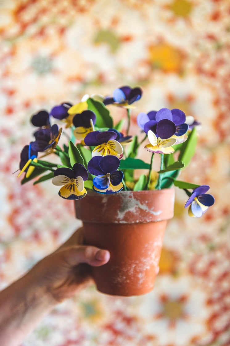 Paper pansies in a distressed terracotta pot against a blurred warm background.