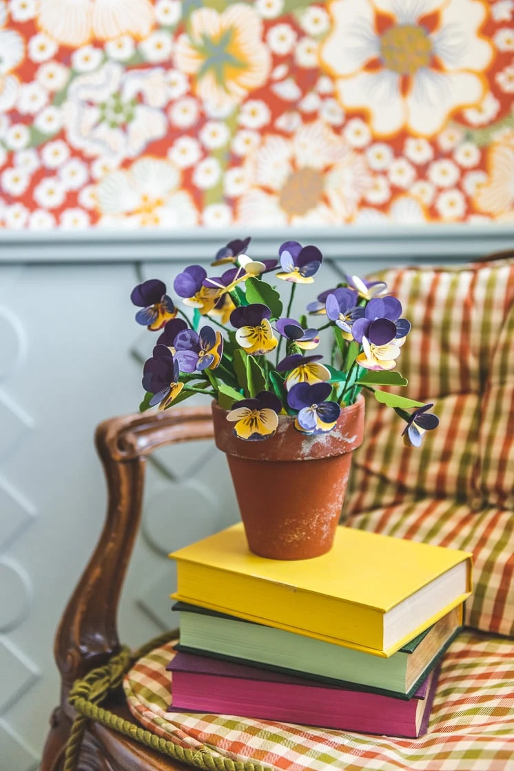 Paper pansies in a distressed terracotta planter. They're placed on a stack of colorful books on a chair. In the background, you can see some red floral wallpaper and blue wainscoting.