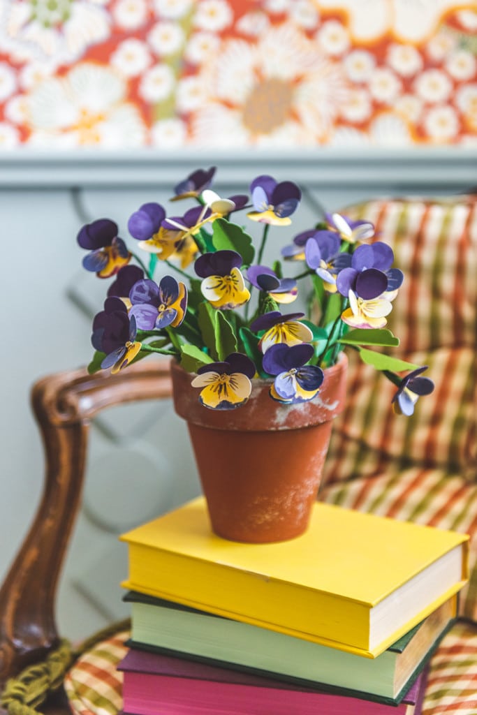 Paper pansies in a distressed terracotta planter. They're placed on a stack of colorful books on a chair. In the background, you can see some red floral wallpaper and blue wainscoting.