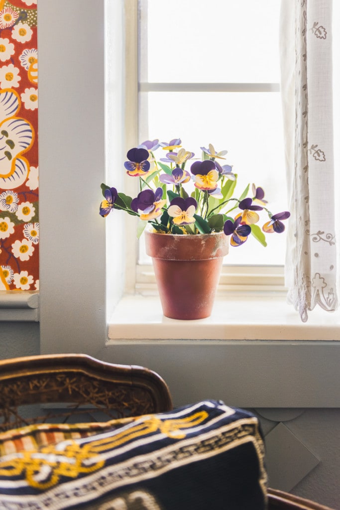 Paper pansies on a windowsill. There's a white lacy curtain next to them, and red floral wallpaper on the other.