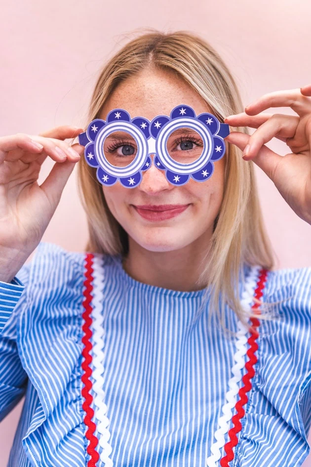 A blonde woman in a blue striped dress with red and white rick rack wears blue paper glasses with white accents in the shape of round flowers. She's standing against a blush pink background. 