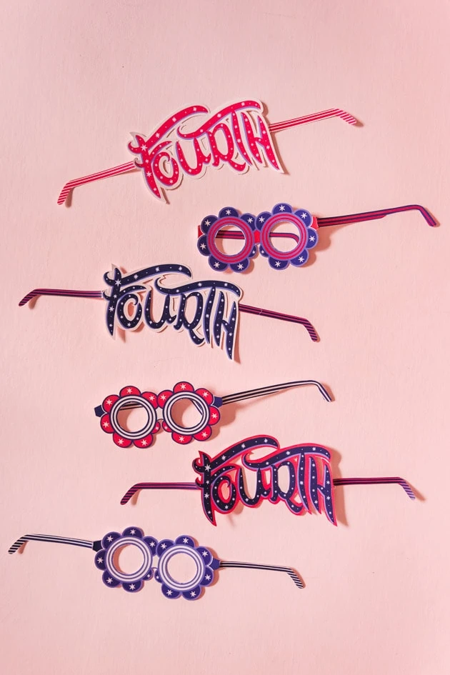 various paper glasses, some with a round floral shape and some reading "fourth," in red and blue with white accents. They're on a blush pink background.