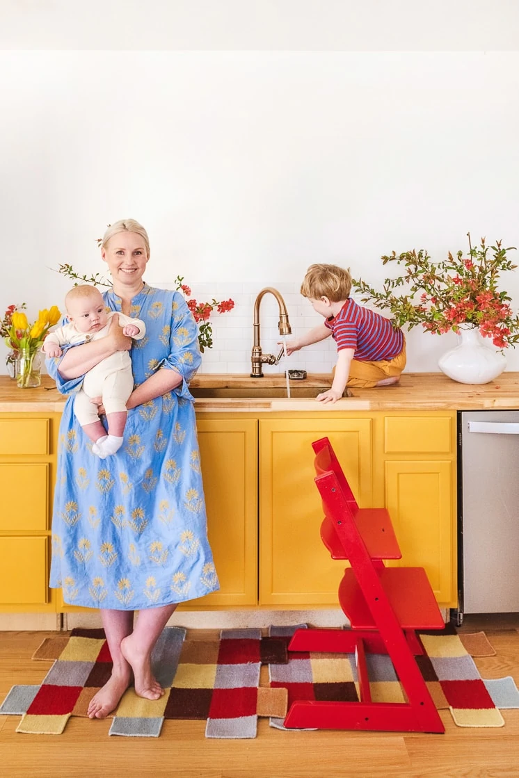 Brittany is wearing a blue dress and holding a baby. She's standing in front of a yellow cabinet