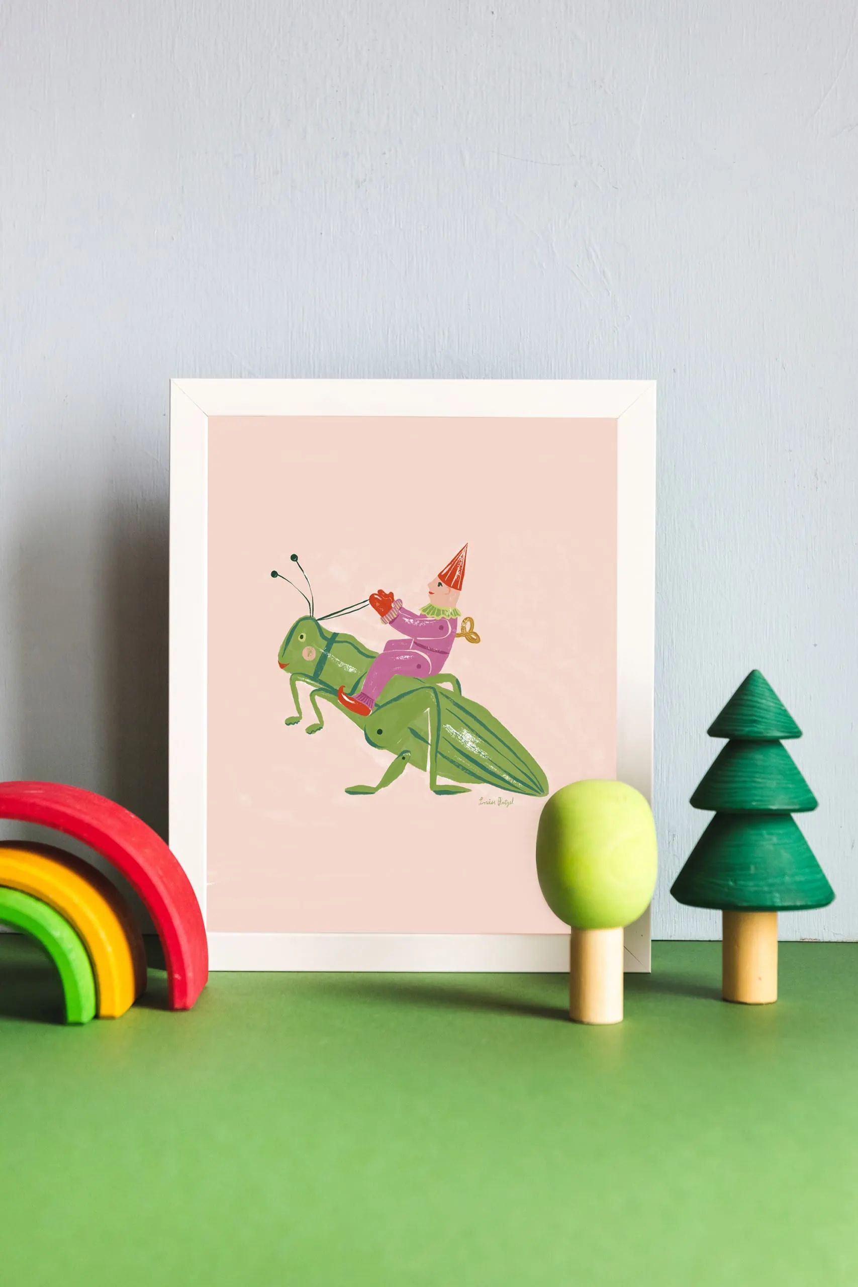 A print of Louise Pretzel's Grasshopper leaning against a light blue wall with wooden rainbow and tree toys in front of it.