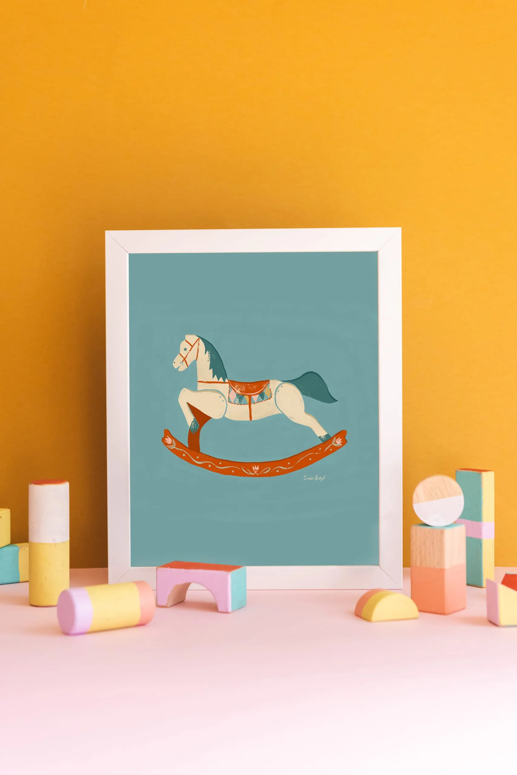 A print of Louise Pretzel's Rocking Horse leaning against a gold wall with pastel wooden blocks in front of it. 