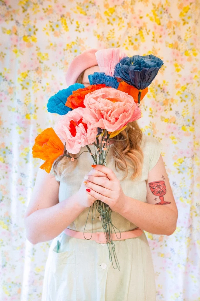 Louise Pretzel stands against a floral backdrop wearing a pink beret and a dress. She's holding paper flowers in front of her face.