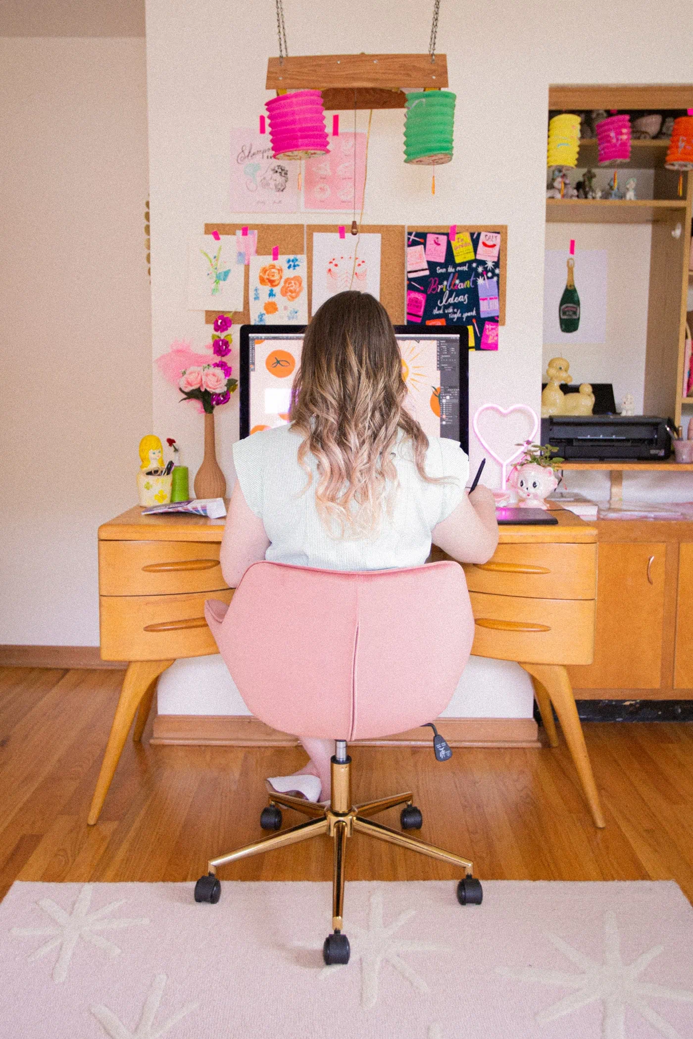Shot of Louise sitting at her desk from behind. The image is symmetrically composed and a corkboard with colorful notes and drawings hangs against the wall.