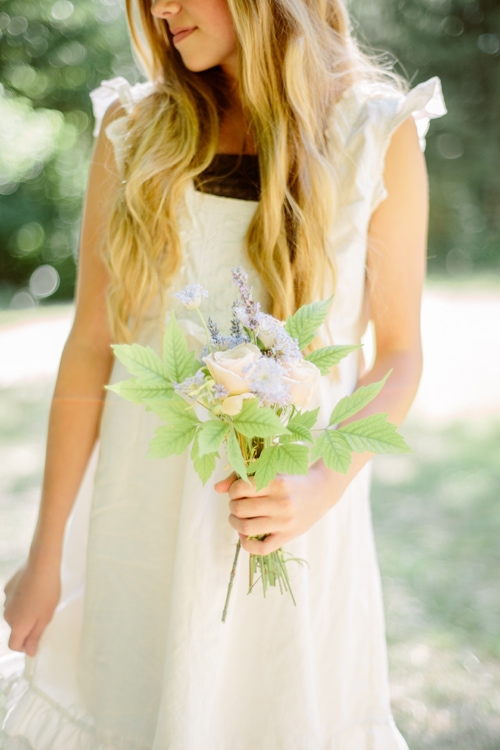 A blonde woman in a white dress holds a small bouquet.