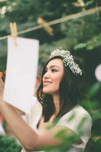 A woman in white wearing a white floral crown hangs up a picture on a clothesline