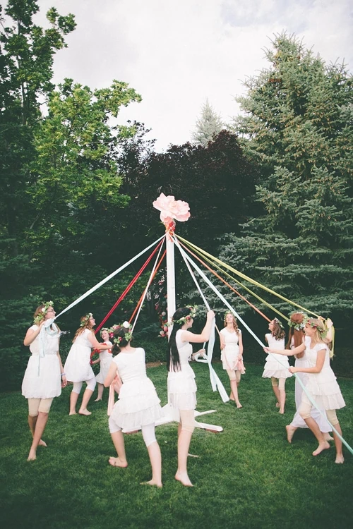 Women in white dance around a maypole in a park filled with trees. It's dusk.