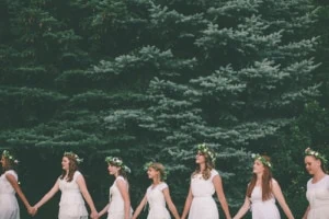 Women in white wearing flower crowns hold hands and walk in a line in front of some pine trees.