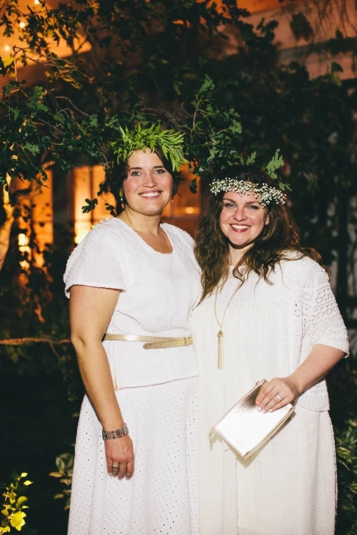 Two women in white wearing floral crowns smile at the camera. It's night and the background is distantly lit with warm light. 