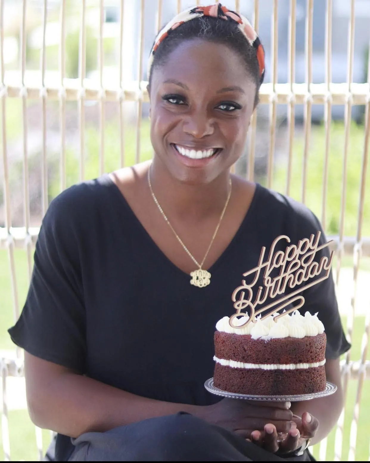 Sheryl Garner holds a red velvet cake and smiles at the camera. She's wearing a black top and a gold necklace.