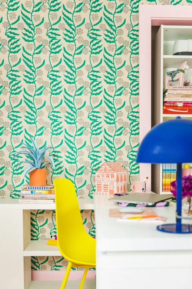 A brightly colored office. There's green floral wallpaper, a yellow chair, white tables and shelves, pink-painted doorframes, a blue modern lamp, an orange paper house, ad office supplies.